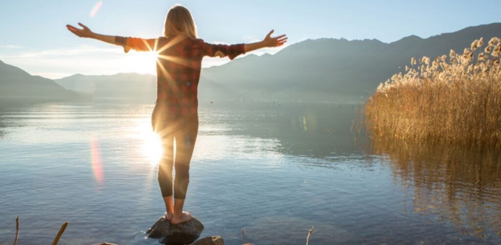 therapie-tegernsee-schnuppermeditation2 Frau steht mit ausgebreiteten Armen am Ufer eines Sees mit Bergpanorama und schaut in die untergehende Sonne